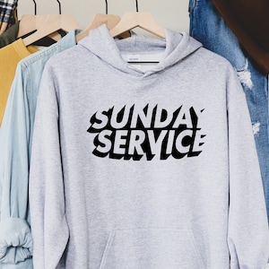 May include: A light gray hoodie with the words "SUNDAY SERVICE" in bold, black, stylized lettering. The hoodie has a hood and is hanging on a wooden hanger. Other clothing items are visible in the background.