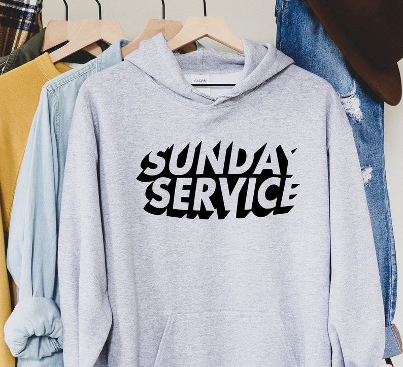 May include: A light gray hoodie with the words "SUNDAY SERVICE" in bold, black, stylized lettering. The hoodie has a hood and is hanging on a wooden hanger. Other clothing items are visible in the background.