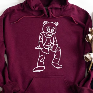 Pu&ograve; includere: Una felpa con cappuccio bordeaux con un'illustrazione a contorno bianco di un orso. L'orso indossa un costume ed &egrave; seduto. La felpa ha un cordino e una tasca frontale.