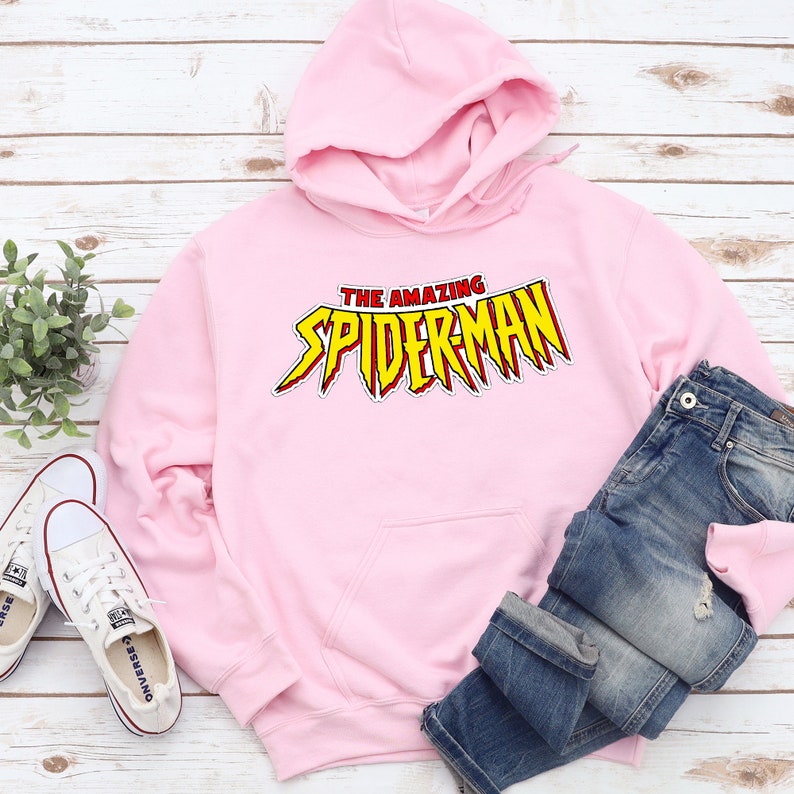 May include: A light pink hoodie with the words "THE AMAZING SPIDERMAN" in a red and yellow comic book style. The hoodie is paired with blue jeans and white sneakers on a light wood background.