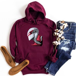 May include: Burgundy hoodie with a graphic of a futuristic helmet design. The helmet is white, gray, and red, with the number "2099" printed on it. The hoodie is paired with blue jeans and brown flats.