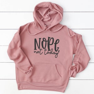 May include: A dusky pink hoodie with a drawstring hood and a front pocket. The hoodie displays the phrase "Nope, not today!" in black script across the front. The hoodie is made of a soft material and is displayed on a white wooden surface.