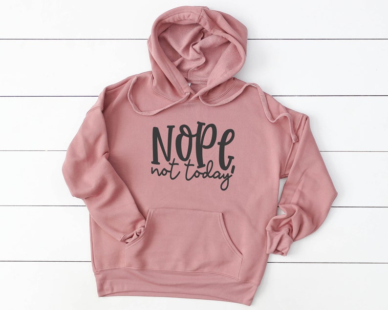 May include: A dusky pink hoodie with a drawstring hood and a front pocket. The hoodie displays the phrase "Nope, not today!" in black script across the front. The hoodie is made of a soft material and is displayed on a white wooden surface.