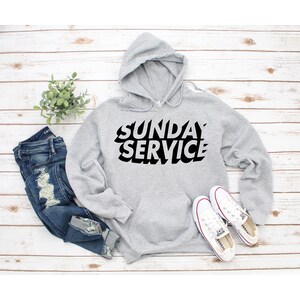 May include: A light gray hoodie with the words "SUNDAY SERVICE" in bold black letters. The hoodie has a front pocket and a hood with a drawstring. White sneakers and blue jeans are also in the image.