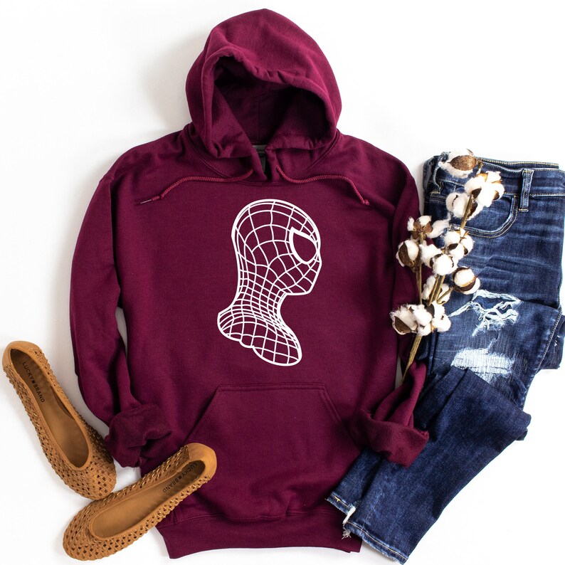 Op de afbeelding: Een bordeauxrode hoodie met een witte afbeelding van een gestileerd spinnenachtig gezicht. De hoodie heeft een capuchon met trekkoord en een voorzak. De afbeelding bevat ook een spijkerbroek en bruine ballerina's.