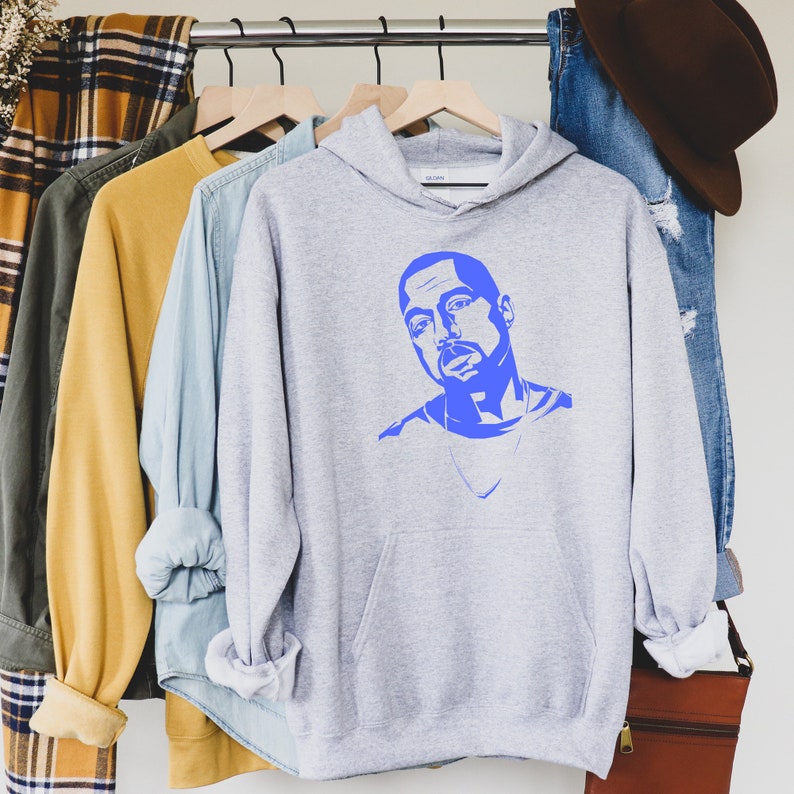 Op de afbeelding: Een lichtgrijze hoodie met een blauwe grafische afbeelding van een persoon. De hoodie heeft een voorzak en een capuchon. Andere kledingstukken, waaronder een geruit overhemd, een gele trui en een spijkeroverhemd, hangen aan een kledingrek.