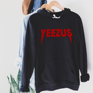 May include: Black hoodie with a red "YEEZUS" graphic. The hoodie has a front pocket and a drawstring hood. The text is in a bold, stylized font. The hoodie is hanging on a wooden hanger.