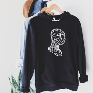 Op de afbeelding: Zwarte hoodie met een witte afbeelding van een gestileerde Spider-Man-kop. Het ontwerp heeft een rasterpatroon, waardoor een 3D-effect ontstaat. De hoodie heeft een voorzak en een wit trekkoord.