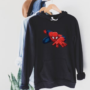 K&ouml;nnte beinhalten: Schwarzer Kapuzenpullover mit einem Spider-Man-Motiv. Der Superheld ist in seinem rot-blauen Anzug dargestellt, mit ausgestreckter Hand. Der Pullover hat eine K&auml;ngurutasche und einen wei&szlig;en Kordelzug. Das Kleidungsst&uuml;ck h&auml;ngt an einem Holzb&uuml;gel.