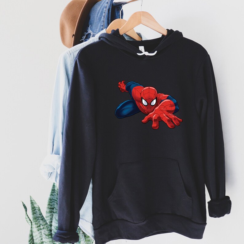 K&ouml;nnte beinhalten: Schwarzer Kapuzenpullover mit einem Spider-Man-Motiv. Der Superheld ist in seinem rot-blauen Anzug dargestellt, mit ausgestreckter Hand. Der Pullover hat eine K&auml;ngurutasche und einen wei&szlig;en Kordelzug. Das Kleidungsst&uuml;ck h&auml;ngt an einem Holzb&uuml;gel.