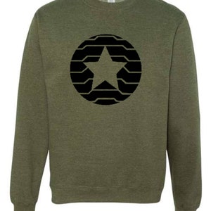 Puede incluir: Sudadera verde oliva con un dise&ntilde;o de estrella negra dentro de un patr&oacute;n circular. La estrella est&aacute; centrada y el c&iacute;rculo est&aacute; compuesto por l&iacute;neas horizontales. La sudadera tiene cuello redondo y mangas largas, adecuada para uso casual.