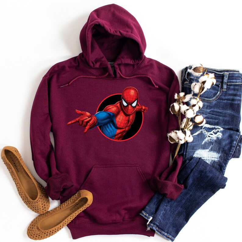 May include: A maroon hooded sweatshirt featuring a graphic of Spider-Man in a black circle. The superhero is depicted in his classic red and blue suit, with his arm extended. The sweatshirt is paired with blue jeans and brown flats.