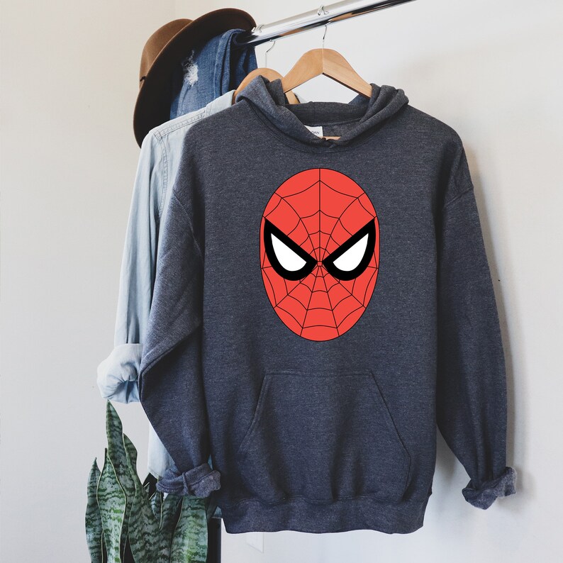 May include: A charcoal gray hooded sweatshirt featuring a large red and black Spider-Man mask design on the front. The sweatshirt has a kangaroo pocket and is hanging on a wooden hanger with other clothing items.