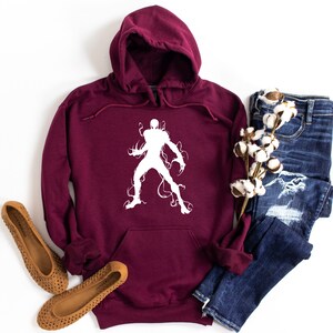 May include: A maroon hooded sweatshirt with a white graphic of a superhero. The sweatshirt has a front pocket and a drawstring hood. The image also shows a pair of blue jeans and brown flats.