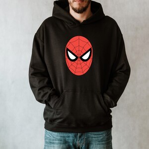 May include: Black hooded sweatshirt featuring a red and black Spider-Man mask design on the front. The graphic is an oval shape with a spiderweb pattern and white eye shapes. The sweatshirt has a front pocket and a drawstring hood.