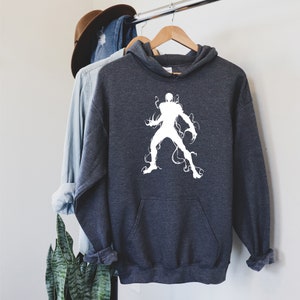 May include: Dark gray hooded sweatshirt with a white graphic of a stylized figure with tendrils. The hoodie has a front pocket and a drawstring hood. A denim shirt and a brown hat hang on a clothing rack behind the hoodie.