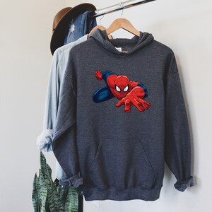 K&ouml;nnte beinhalten: Ein anthrazitfarbener Kapuzenpullover mit einem Spider-Man-Motiv. Der Superheld ist in seinem klassischen rot-blauen Anzug dargestellt, die Hand ausgestreckt. Der Pullover h&auml;ngt an einem Holzkleiderb&uuml;gel.