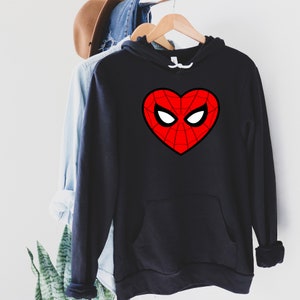 May include: Black hooded sweatshirt with a red heart-shaped design featuring a spiderweb pattern and white eye shapes. The sweatshirt has a front pocket and a white drawstring. The garment is hanging on a wooden hanger.