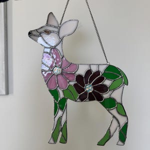 May include: Stained glass deer with a floral design. The deer is white with pink and burgundy flowers, green leaves, and a silver chain for hanging. The deer's body is made of multiple glass pieces.