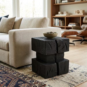 Peut inclure: Une table d'appoint carrée noire faite de blocs de bois empilés. La table a une surface texturée et est placée sur un tapis à motifs à côté d'un canapé beige. Un petit bol rond est posé dessus.