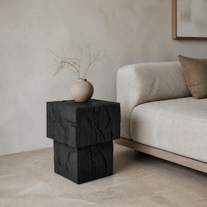 May include: A black, stacked wooden side table with a textured surface. A small, beige vase with dried branches sits on top. The table is next to a beige sofa with a brown cushion.