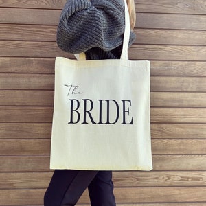 Könnte beinhalten: Cremefarbene Tragetasche mit dem schwarzen Aufdruck "The BRIDE". Die Tasche wird von einer Person getragen, die einen grauen Pullover und eine schwarze Hose trägt. Der Hintergrund ist eine Holzwand.