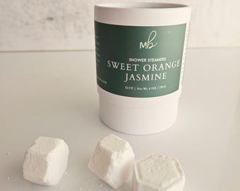 May include: A white cylindrical container with a green label featuring the text "MB SHOWER STEAMERS SWEET ORANGE JASMINE 12 CT | Net Wt. 6 OZ / 170 G". Three small, white, hexagonal-shaped shower steamers are placed in front of the container.