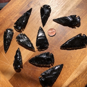 Puede incluir: Un conjunto de puntas de flecha de obsidiana negra, cada una con una punta afilada y formas variadas. Las puntas de flecha se exhiben sobre una superficie de madera, con una moneda para comparar el tamaño. La obsidiana tiene una superficie brillante y reflectante.