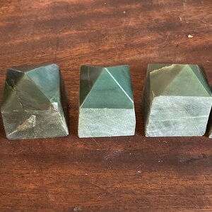 May include: Five green jade pyramid-shaped stones with a faceted design. The stones are displayed on a dark wooden surface. Each stone has a unique pattern and shape, showcasing natural variations in the material.