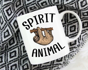 May include: A white ceramic mug featuring a brown sloth hanging from a branch illustration in the center. The text SPIRIT ANIMAL is printed in black capital letters above and below the sloth graphic. The mug has a curved handle on the right side.