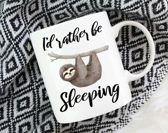 May include: A white ceramic mug featuring a brown sloth hanging from a branch illustration. The text "I'd rather be Sleeping" is printed in black script and sans-serif font above and below the sloth. The mug has a curved handle on the right side.