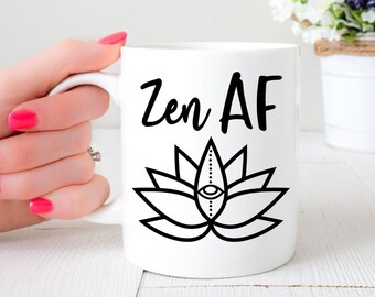 Puede incluir: Una taza de cerámica blanca con un gráfico negro de una flor de loto con un ojo en el centro. Sobre el loto, el texto Zen AF está impreso en una fuente negra, negrita y de estilo manuscrito. La taza tiene un cuerpo cilíndrico y un asa curva.