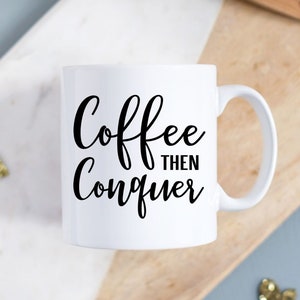 以下が含まれることがあります： 白いセラミック製のコーヒーカップで、黒い文字で「Coffee then Conquer」と書かれています。