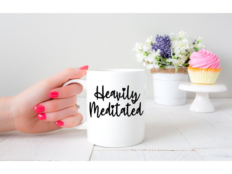 May include: White ceramic coffee mug with black text that reads "Heavily Meditated" with a small heart symbol.