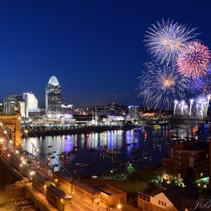 May include: A nighttime cityscape with a bridge and fireworks exploding over a river. The city skyline is lit up with lights, and there are boats on the water.