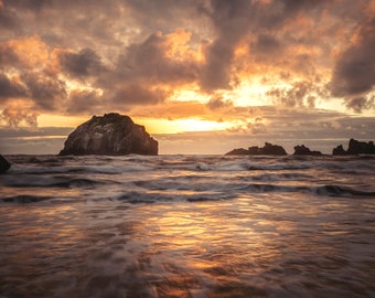 May include: A dramatic sunset over the ocean with a large rock formation in the foreground and smaller rocks in the distance. The sky is a mix of orange, yellow, and purple clouds. The water is a deep blue with whitecaps.