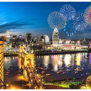 May include: A nighttime view of a city skyline with a bridge and fireworks over a river. The city is lit up with lights and the fireworks are exploding in the sky. There are boats on the river and people gathered on the shore.