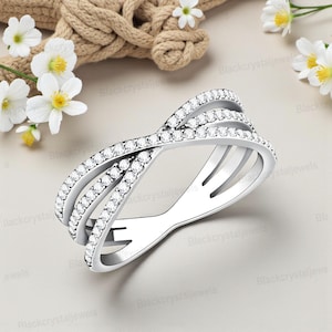 May include: A silver-toned ring with a crisscross design, adorned with numerous small, clear gemstones. The ring features three bands that intersect, creating an open, airy look. The background includes white flowers and a rope.