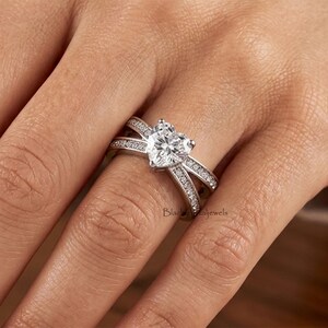 May include: A silver-toned ring featuring a central heart-shaped clear gemstone. The band has a crossover design and is encrusted with small, round, clear stones along both sides. The text BlackPetaljewels is visible on the ring band.