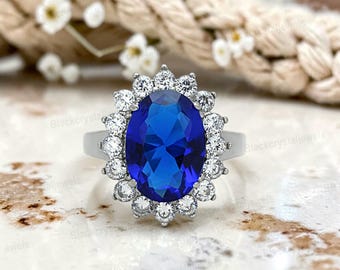 May include: A silver-toned ring featuring a large, oval-cut, deep blue center stone surrounded by a halo of fourteen small, round, clear, sparkling stones. The band is smooth and metallic, providing a clean contrast to the vibrant blue gemstone.