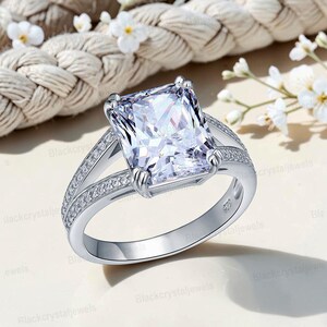 May include: A silver ring with a large, square-cut, clear gemstone as the centerpiece. The band splits into two parallel rows, each lined with smaller, clear stones. The ring is set against a neutral background with white flowers.