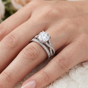 May include: A silver-toned engagement ring set featuring a large, round, brilliant-cut center stone held by four prongs. The band is composed of two intertwined, crisscrossing shanks encrusted with small, clear pavé-set stones throughout.