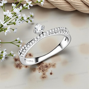 May include: A silver-toned ring featuring a central round-cut clear stone set in a four-prong mount. The band has a split-shank design adorned with small pavé-set clear stones along both sides. The interior of the band is engraved with the text TK316.