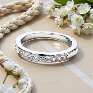 Puede incluir: Un anillo de plata con una fila de piedras claras de corte cuadrado. El anillo se presenta sobre un fondo neutro con flores blancas y una cuerda. El anillo es una joya.
