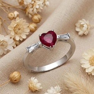 May include: A silver-toned ring featuring a central heart-shaped deep red gemstone flanked by two clear baguette-cut stones on either side. The band is smooth and metallic, and the text Blackcrystaljewels is printed near the base of the ring.