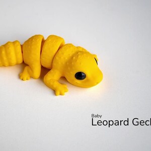 May include: A yellow, segmented, 3D printed toy model of a baby leopard gecko. The toy has large black eyes and a curled tail.