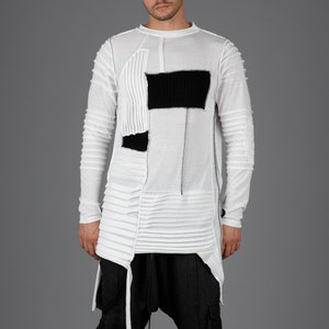 Può includere: Una maglia bianca a maniche lunghe con toppe nere e un design a pieghe unico, abbinata a pantaloni grigio scuro a cavallo basso e sandali neri. L'outfit ha un'estetica avant-garde e moderna.