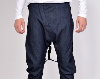 May include: Dark blue denim pants with a unique, draped design. The pants have a high waist and a relaxed fit through the hips, tapering to the ankles. The model is wearing black sneakers with white soles. The pants are made of a dark wash denim.