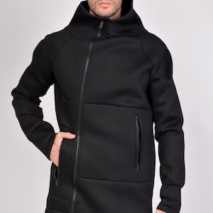 May include: Black hooded jacket with a diagonal zipper closure and multiple zippered pockets. The jacket has a high collar and a textured surface. The model is wearing a white shirt underneath and black pants.