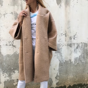 Może przedstawiać: Długi, brązowy płaszcz wełniany z szerokim kołnierzem i dużymi kieszeniami. Płaszcz jest rozpięty, a rękawy podwinięte. Osoba nosząca płaszcz stoi przed szarą ścianą.